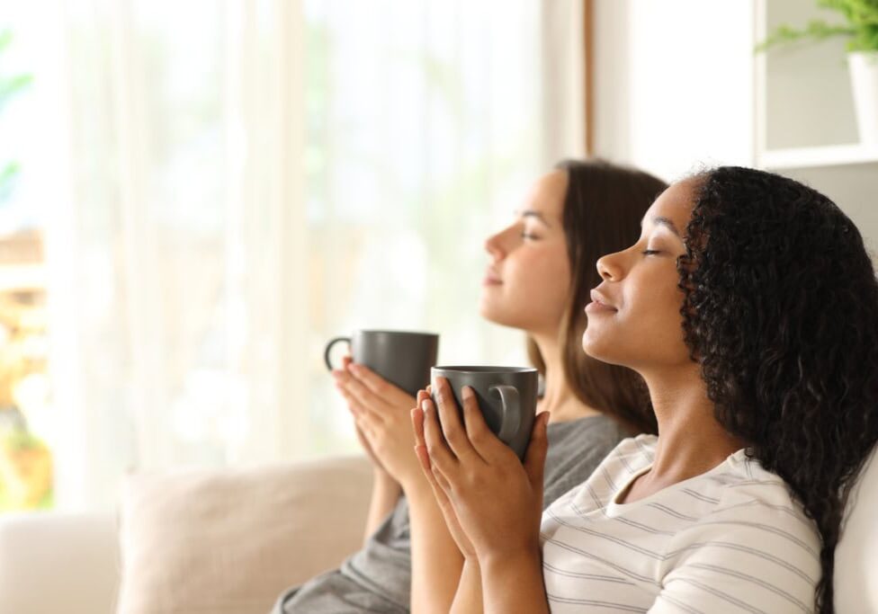 Side view portrait of two interracial roommates relaxing drinking coffee sitting on a couch at home
